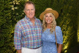 "Debby Clarke Belichick and Bill Belichick posing together at a public event, smiling for the camera while standing in front of an elegant green backdrop. The image highlights the former couple during a social appearance before their divorce."