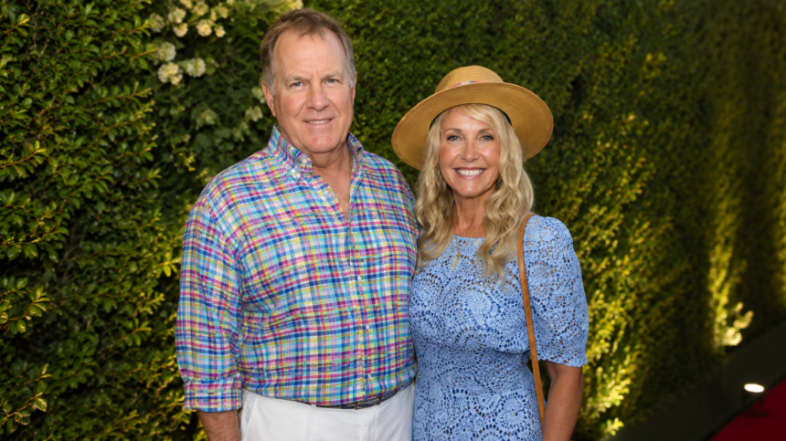 "Debby Clarke Belichick and Bill Belichick posing together at a public event, smiling for the camera while standing in front of an elegant green backdrop. The image highlights the former couple during a social appearance before their divorce."